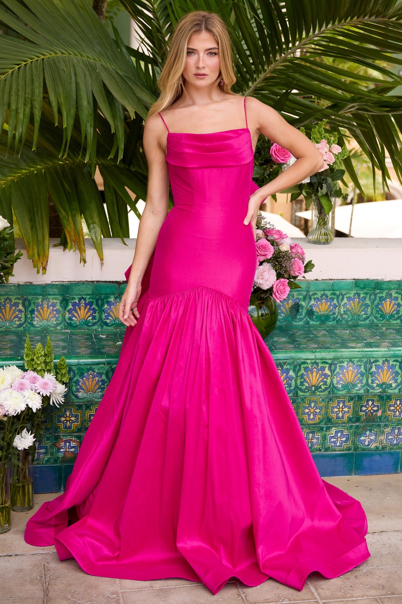 A woman in the Ava Presley 26422 Drop-Waist Mermaid Prom Gown in magenta stands before lush tropical plants and a vibrant tiled wall, surrounded by assorted flowers.